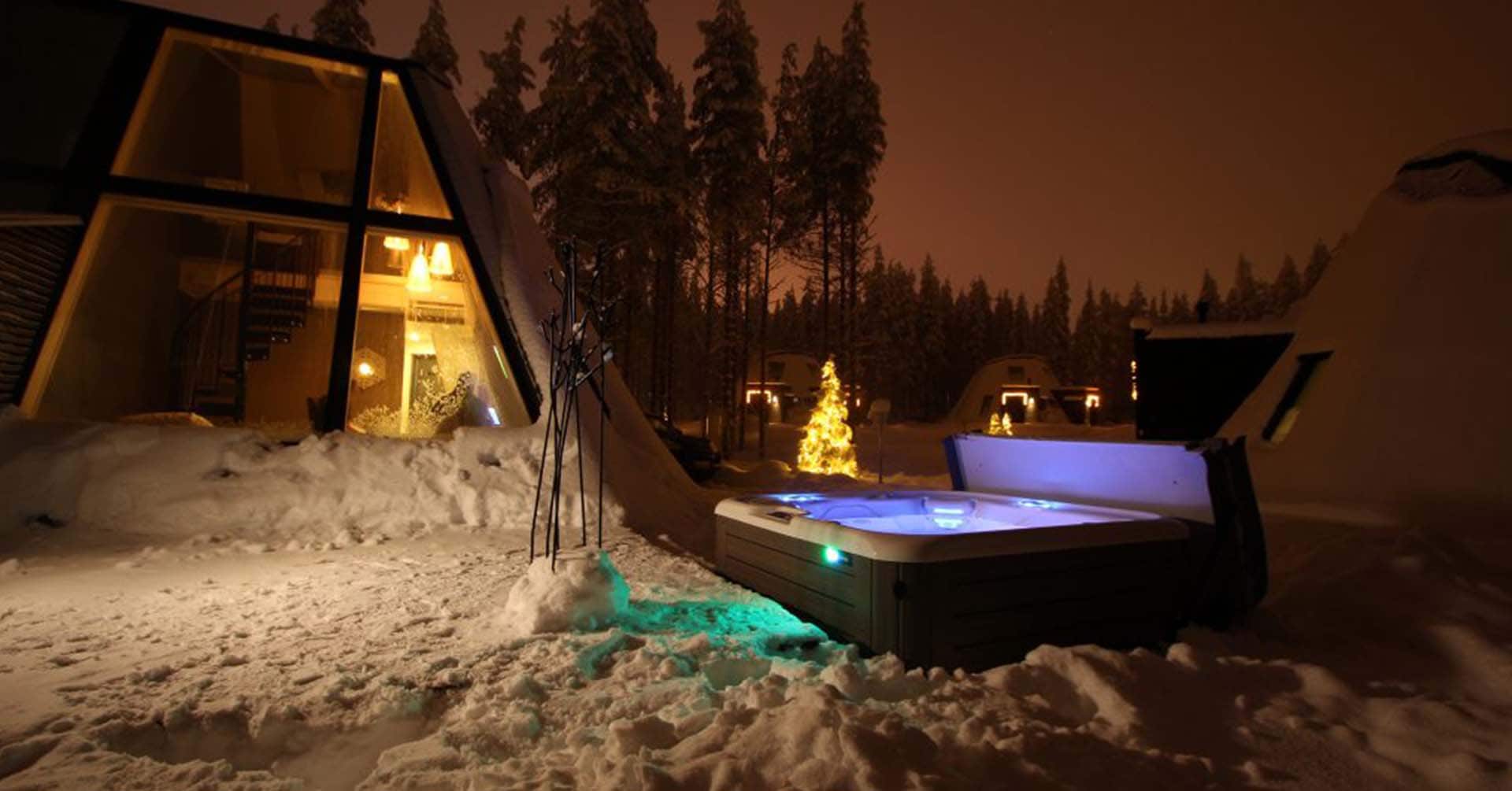 HotSpring HighLife Glass Resort Rovaniemi Ulkoporeallas valot päällä iltahämärässä lumessa pienten erikoisen muotoisten rakennusten edessä.